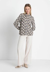 Langärmlige Bluse mit gemusterten, kreisförmigen Designs in Beige und Schwarz, kombiniert mit weißen weiten Hosen und hellbeigen, hohen Schuhen.