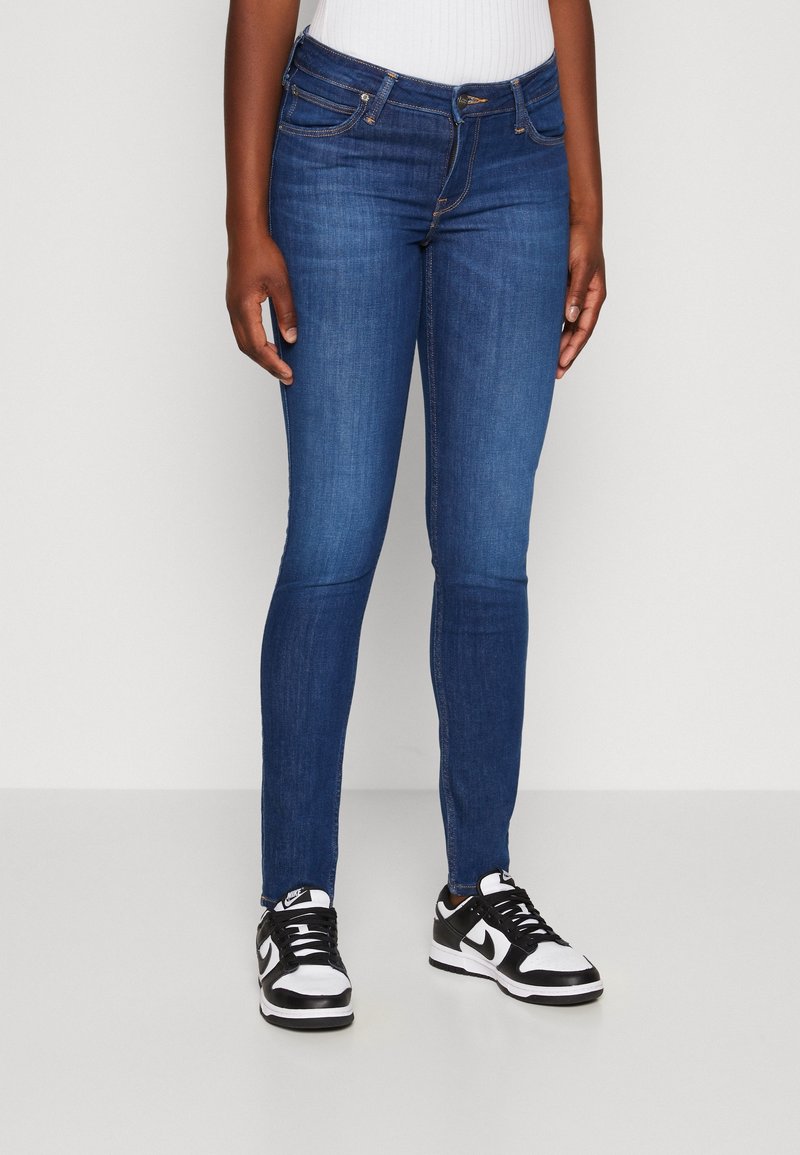 Jean skinny bleu foncé en denim, comprenant une taille mi-haute, cinq poches et des détails de coutures subtiles, associé à des baskets noires et blanches.