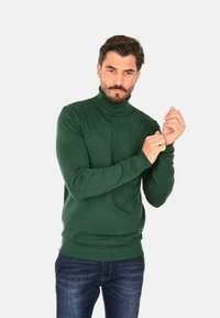 Maglione verde a collo alto realizzato in morbido tessuto a maglia, con maniche lunghe e polsini a coste. Indossato con jeans blu scuri.