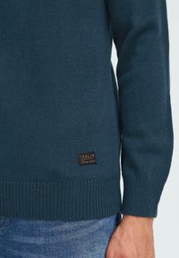 Donkerblauwe gebreide trui met ribgebreide manchetten en zoom, een gestructureerd oppervlak en een kleine bruine leren logo patch aan de onderzijde.