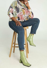 Top de manga larga con estampado floral de hojas multicolores; vaqueros oscuros ajustados; botas verdes brillantes hasta el tobillo con tacón y punta cuadrada.