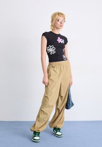 Sort tætsiddende T-shirt med farverige blomsterprint, kombineret med løse beige bukser. Modellen har grønne sneakers på og holder en denimjakke.