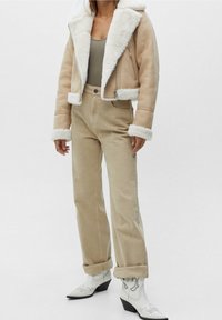 Beige shearling bomberjack met witte nepbont afwerking, gecombineerd met beige hoge taille broek en witte enkellaarsjes met zwarte accenten.