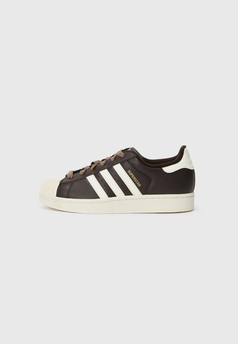 Sneaker Adidas Superstar low-top in pelle marrone scuro con punta a conchiglia crema, strisce bianche e scritta "Superstar" sul lato.