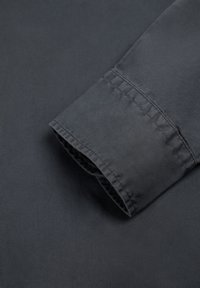 Mørkegrå skjortemanchet med synlige syninger og glat stoftekstur på flad overflade.