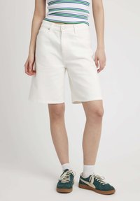 Shorts en denim blanc avec une coupe droite, cinq poches et fermeture à bouton à l'avant. Associés à des baskets teal et des chaussettes blanches.