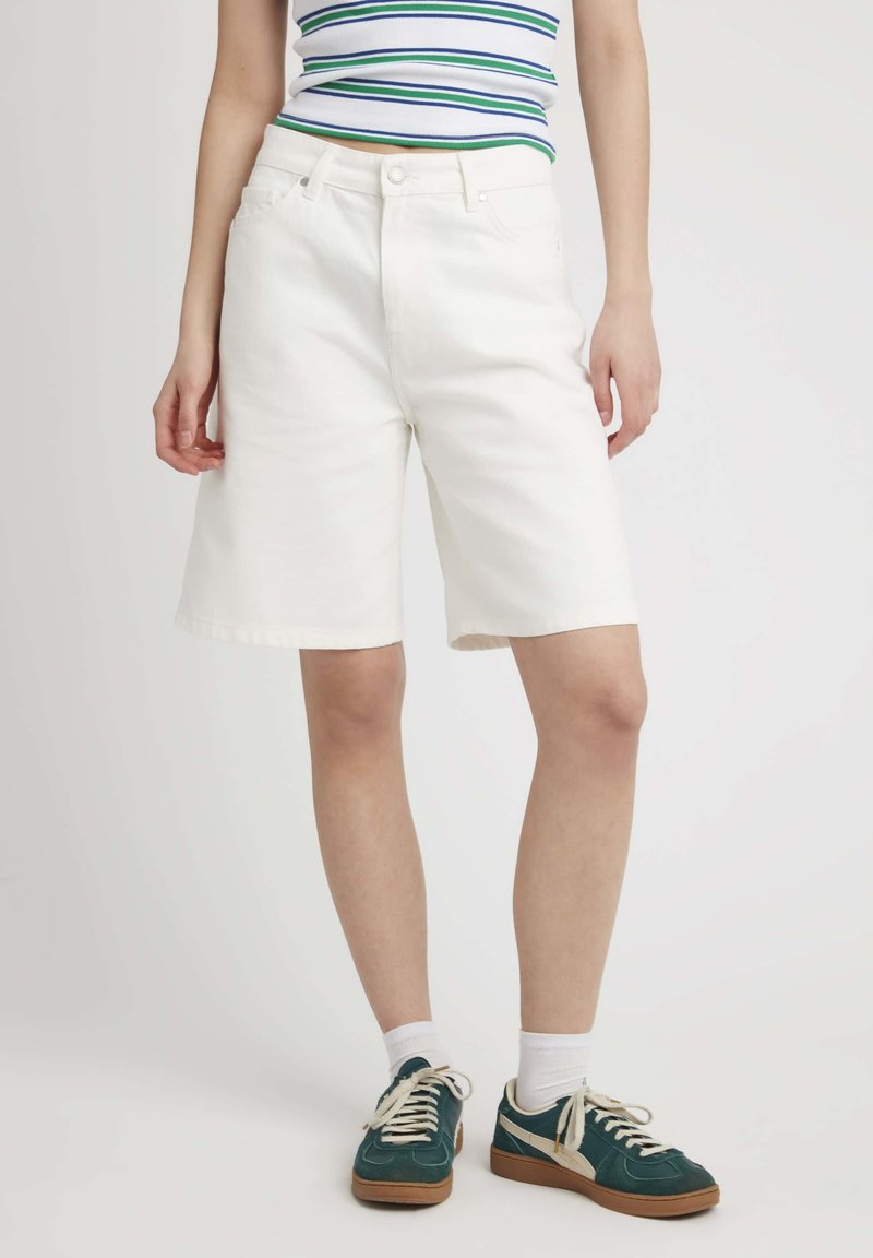 Shorts en denim blanc avec une coupe droite, cinq poches et fermeture à bouton à l'avant. Associés à des baskets teal et des chaussettes blanches.