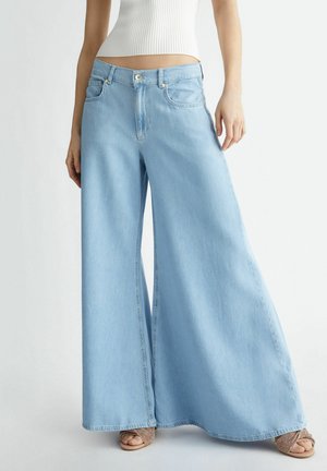 Persoon draagt lichtblauwe jeans met wijde pijpen, een wit kort geribd tanktopje en opengewerkte sandalen met open teen, staand tegen een effen achtergrond.