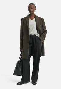 Brauner Wollmantel mit aufgeschlagenem Revers, graues V-Ausschnitt-Shirt, schwarze Hose mit elastischem Bund und eine strukturierte schwarze Tragetasche. Schwarze Slipper.
