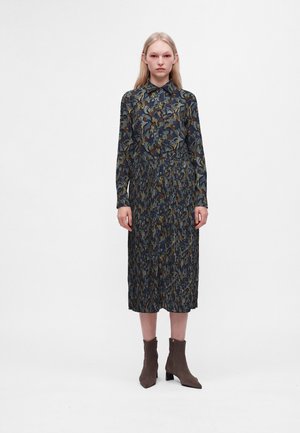 Jeune femme portant une robe midi à manches longues à imprimé floral foncé avec des bottines marron, debout devant un fond blanc uni.