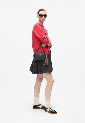Sac à main noir texturé avec une fermeture éclair argentée et des étiquettes rose et marron, associé à un sweat-shirt rouge et une jupe plissée noire.