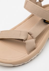 Beige sandal featuring two wide fabric straps, textured sole, and curved footbed design. Adjustable buckle and logo detail on the strap.
