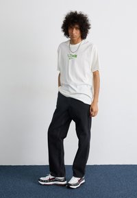 Hvid bomulds t-shirt med grøn grafisk print, kombineret med sorte denim bukser og hvide sneakers med røde og sorte accenter.