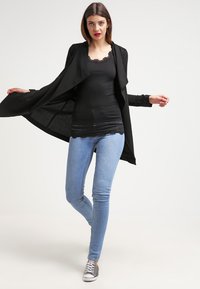 Zwarte open-front cardigan met een vloeiende uitstraling, gedragen over een zwart met kant afgewerkte tanktop en lichtblauwe skinny jeans. Grijze sneakers worden gedragen.