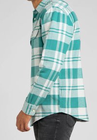 Chemise en flanelle à carreaux turquoise et blanche avec manches longues, poignets boutonnés et ourlet incurvé. Dispose d'une poche poitrine et d'un tissu texturé.