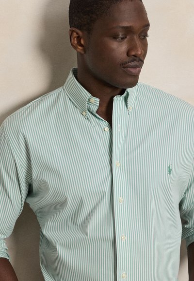 Mężczyzna w zielono-białej paskowanej koszuli na guziki z małym logo na piersi, lekko patrzący w bok na jednolitym tle.