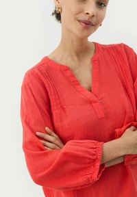Czerwona lniana bluzka z dekoltem w kształcie litery V, zebranymi mankietami i subtelnymi kantami. Posiada luźny krój i miękką teksturę.