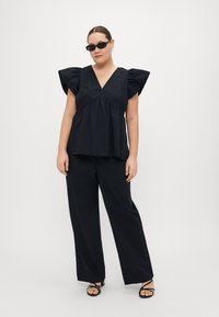 Black relaxed-fit top with voluminous ruffled sleeves, paired with wide-leg black trousers and strappy black sandals. Minimalist design.