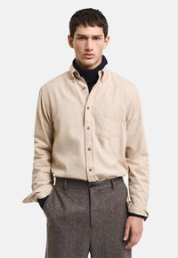 Camisa de botón de color beige claro con un bolsillo en el pecho, llevada sobre un cuello de tortuga azul marino, combinada con pantalones grises texturizados.