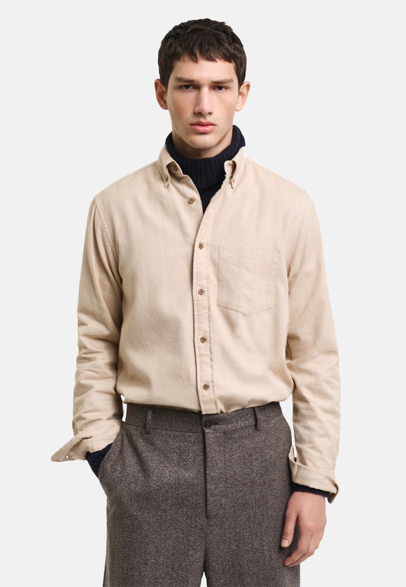 Camisa de botón de color beige claro con un bolsillo en el pecho, llevada sobre un cuello de tortuga azul marino, combinada con pantalones grises texturizados.