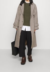 Cappotto lungo a quadri beige e marrone, maglione in maglia oliva, pantaloni larghi neri e stivaletti marroni con tacco a blocco. Borsa tote nera accanto.