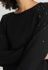Haut noir à manches longues avec de petites clous métalliques sur les épaules et le haut des bras, porté par une personne aux cheveux bouclés et aux bras croisés.