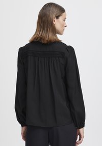 Svart långärmad blus med avslappnad passform, plisserad bak och texturerat okdetalj. Mjuk tyg med rynkade manschetter.