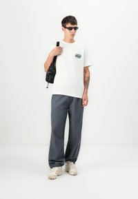 Witte T-shirt met een groene cirkelvormige logo, grijze losse broek met elastische boorden, zwarte schoudertas en beige sneakers.