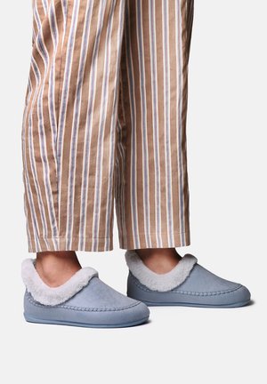 Pieds portant des chaussons bleu clair à enfiler avec doublure en fourrure blanche, associés à un pantalon beige et blanc rayé.