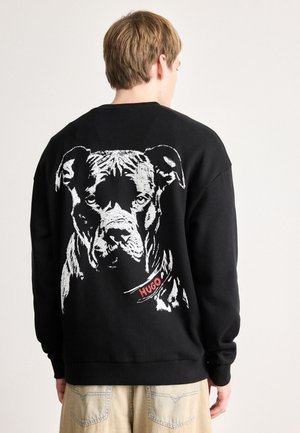 Junge Person, von hinten gesehen, trägt einen schwarzen Pullover mit einem großen weißen Hundegesicht und "HUGO" am Kragen, das auf dem Rücken aufgedruckt ist.