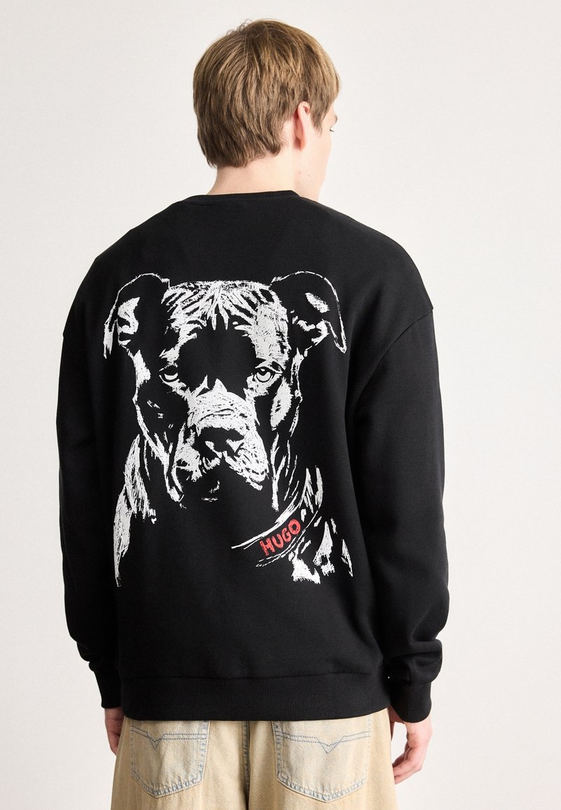 Jeune personne de dos portant un sweat-shirt noir avec un grand visage de chien blanc et "HUGO" sur le col imprimés dans le dos.