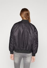 Černá bomber bunda s hladkou, lesklou texturou, pružnými manžetami a pasem. Obsahuje zipovou kapsu na levém rukávu.