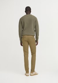 Olivgrüner Strickpullover mit rundem Ausschnitt, kombiniert mit beigen Chinos. Beigefarbene Sneaker mit drei Streifen. Rückansicht des Outfits.