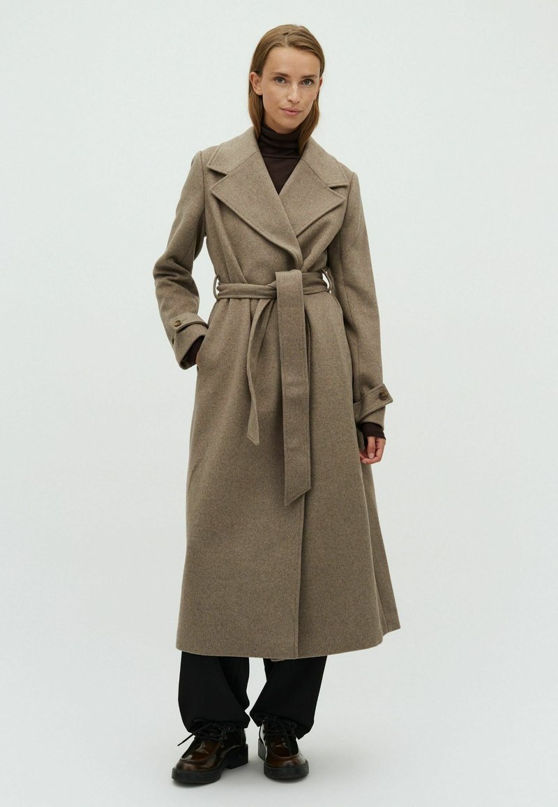 Beige wollen trenchcoat met een brede revers, een ceintuur op de taille en lange mouwen, gecombineerd met zwarte broek en laarzen.