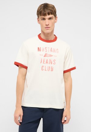 Junger Mann trägt weißes T-Shirt mit rotem Rand und verblasstem "Mustang Jeans Club"-Schriftzug, kombiniert mit dunkelblauer Hose, steht vor schlichten Hintergrund.