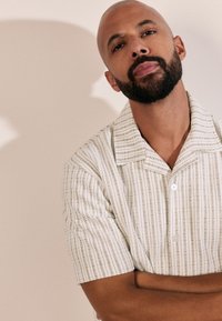 Camicia a maniche corte in crema chiaro con righe verticali verdi, tessuto strutturato e colletto con grandi bottoni.