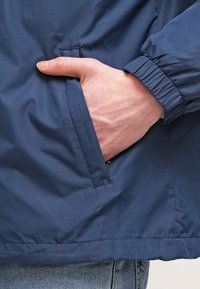 Ruka zasunutá v boční kapse modré bundy s elastickým manžetovým nástřelem přes džíny.