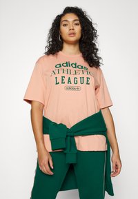 Persikų spalvos medvilninis marškinėliai su žaliais užrašais "adidas ATHLETIC LEAGUE", derinami su žaliais džinsais ir žaliai surištu megztiniu aplink liemenį.