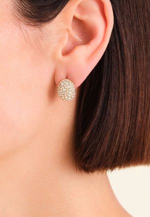 Gold-tone stud earrings with a textured oval shape, adorned with numerous small clear crystals for added sparkle, set against short, dark hair.