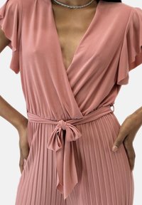 En rosa plisserad jumpsuit med en djup V-ringning, volangärmar och ett knytbälte. Slät tyg med en mjuk textur och en följsam design.