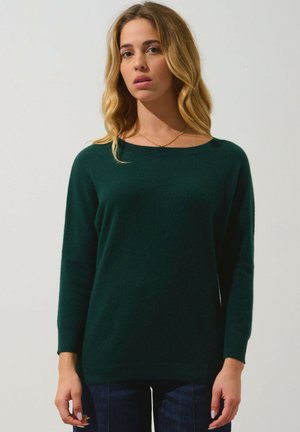 Pull en tricot vert foncé avec un col rond, des manches longues et des poignets côtelés, associé à un jean bleu. Texture lisse, design décontracté.