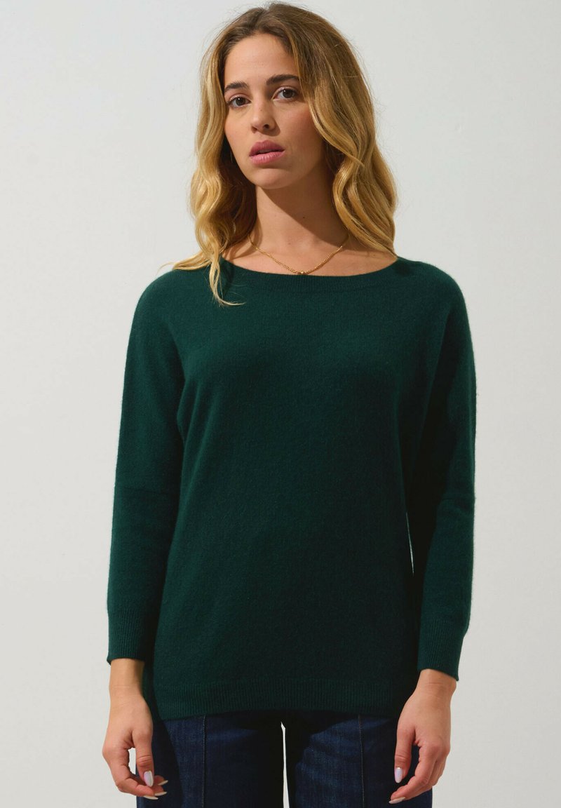 Pull en tricot vert foncé avec un col rond, des manches longues et des poignets côtelés, associé à un jean bleu. Texture lisse, design décontracté.