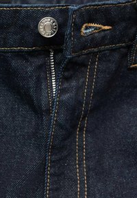 Tissu en denim bleu foncé avec des coutures visibles, bouton en métal et fermeture éclair. Vue en gros plan de la taille et de la braguette.