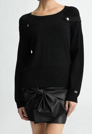Black long-sleeve sweater with buttoned shoulder straps paired with a black leather mini skirt featuring a large knotted bow.
