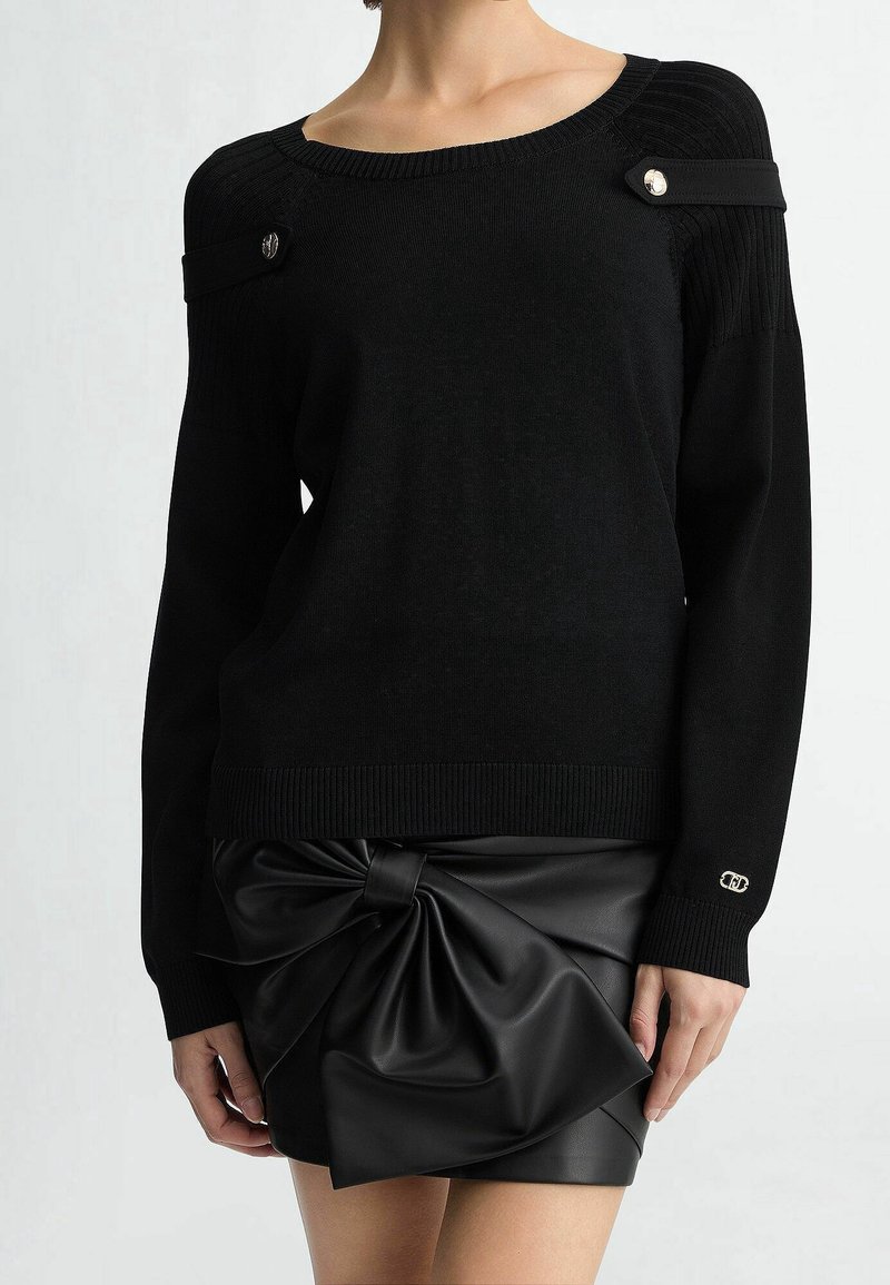Pull noir à manches longues avec bretelles boutonnées aux épaules, assorti à une mini-jupe en cuir noire ornée d'un grand nœud noué.
