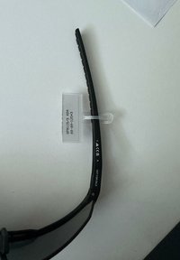 Braccio di occhiali da sole neri con testo bianco "NON IN VENDITA" e marcatura CE, tenuto da un'etichetta di plastica trasparente su una superficie bianca.