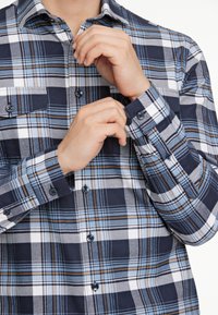 Camicia a quadri blu e bianca con dettagli marroni, caratterizzata da una chiusura a bottoni e due tasche sul petto, realizzata in morbido cotone.