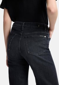 Jeans in denim scuro con vita alta, cinque tasche e dettagli di cucitura sottili. Il tessuto ha una texture liscia e leggermente lucida.