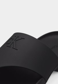 Μαύρο σανδάλι με φαρδιά λουρί που φέρει ανάγλυφο λογότυπο "cK" σε ανάγλυφα μαύρα γράμματα.