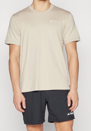 Mężczyzna noszący beżowy t-shirt Terrex z krótkim rękawem i czarne szorty Terrex, stojący na jednolitym białym tle.